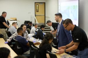 A group of people in a classroom participate in a hands-on workshop, with instructors assisting attendees at tables.