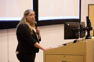 A woman holding a microphone stands at a podium with computer monitors, speaking in front of a projection screen in a lecture hall.