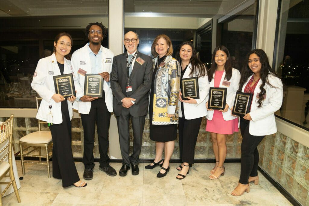 Group shot of five WesternU student scholars and the President and Provost of WesternU. The students are holding plaques. 