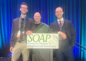 Three men standing together behind a podium with a sign that reads "SOAP Society for Obstetric Anesthesia and Perinatology." The background is a blue curtain.