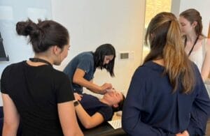 A person receives a medical procedure while lying on a table. Three individuals observe attentively. All are in a well-lit indoor setting.