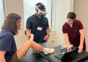 Three men in a medical setting: one sitting with a raised leg, another examining it while wearing a head-mounted device, and a third operating a tablet.