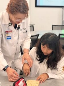 An adult in a white lab coat assists a young girl, also in a lab coat, with a medical task using tools on a table.