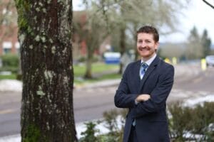 A man in a suit with a striped tie stands with his arms crossed in an outdoor setting with trees and a road in the background.
