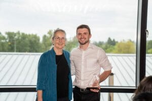 A woman and a man stand together indoors in front of a large window with a view of trees. The man is holding a clear triangular-shaped award. Both are smiling at the camera.
