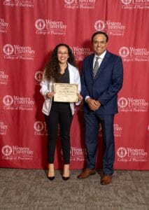Two people stand in front of a red "Western University College of Pharmacy" backdrop. One holds a scholarship certificate.