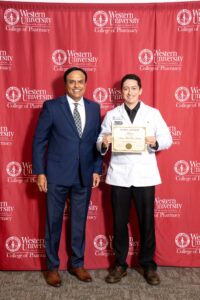 Two individuals stand in front of a red Western University College of Pharmacy backdrop. One is in a suit and the other holds a certificate while wearing a white coat.