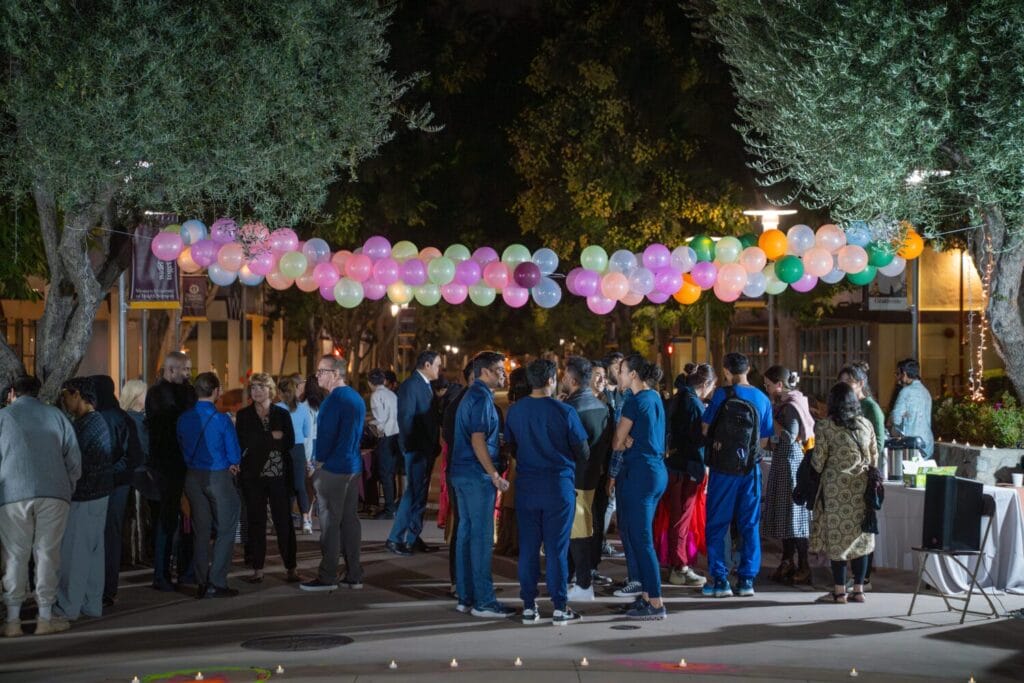 A group of people gather outdoors at night under a string of colorful balloons, surrounded by trees and lights.