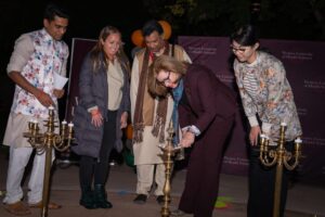 A group of people, some in traditional attire, gather around a woman lighting a ceremonial lamp at an event.
