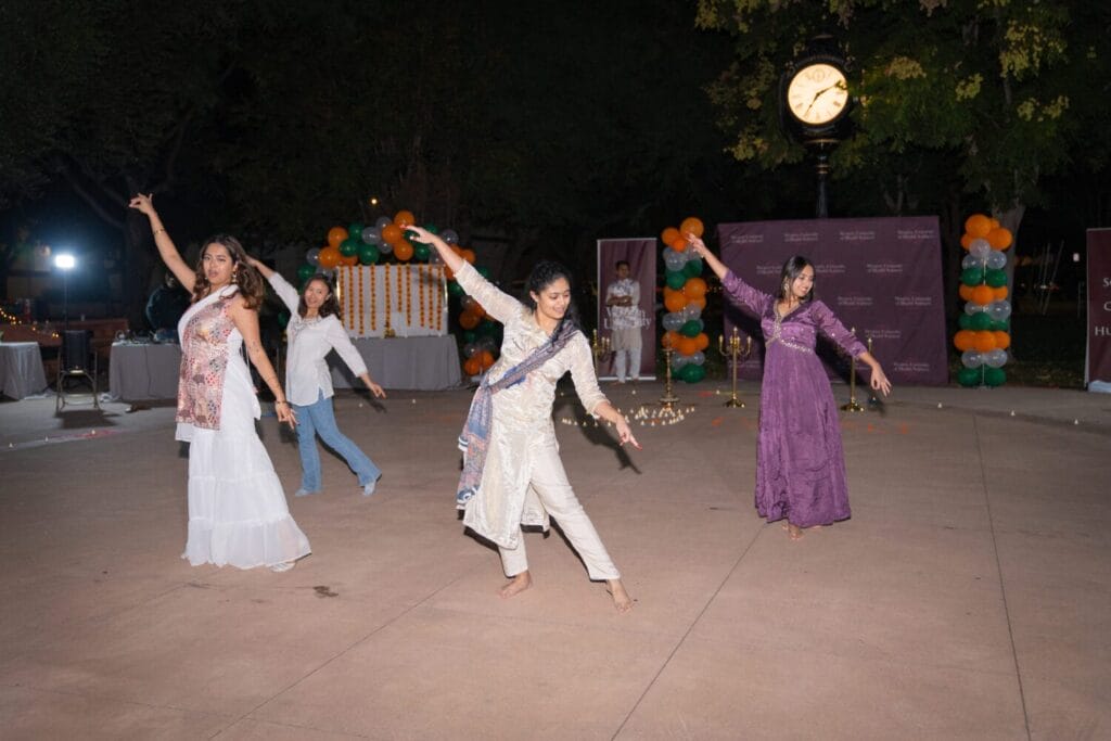 Four people dancing outdoors, each in traditional attire. Decor includes balloons and a clock in the background.