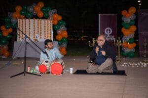 Two people seated on stage with microphones at a cultural event, featuring colorful balloons and a backdrop. One person plays the tabla while the other speaks.