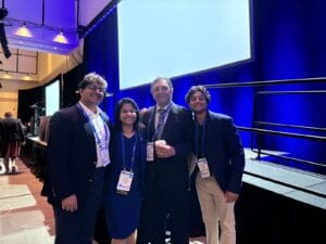 Four people in business attire stand smiling in front of a stage with blue lighting.
