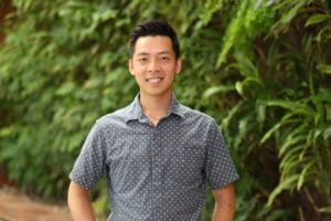 Person standing outdoors, smiling, wearing a gray patterned shirt, with a green leafy background.