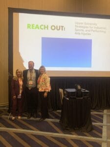 Three people stand in front of a presentation screen titled "Reach Out: Upper Extremity Strategies for Industrial, Sports, and Performing Arts Injuries.