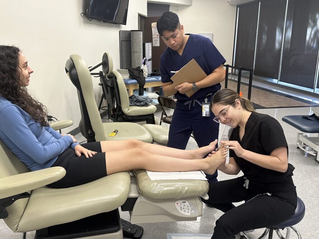 A woman sits on a chair receiving a foot exam from a healthcare worker while another person observes and takes notes in a medical setting.