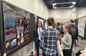 People stand and converse in front of scientific research posters at a conference poster session in a large indoor venue.