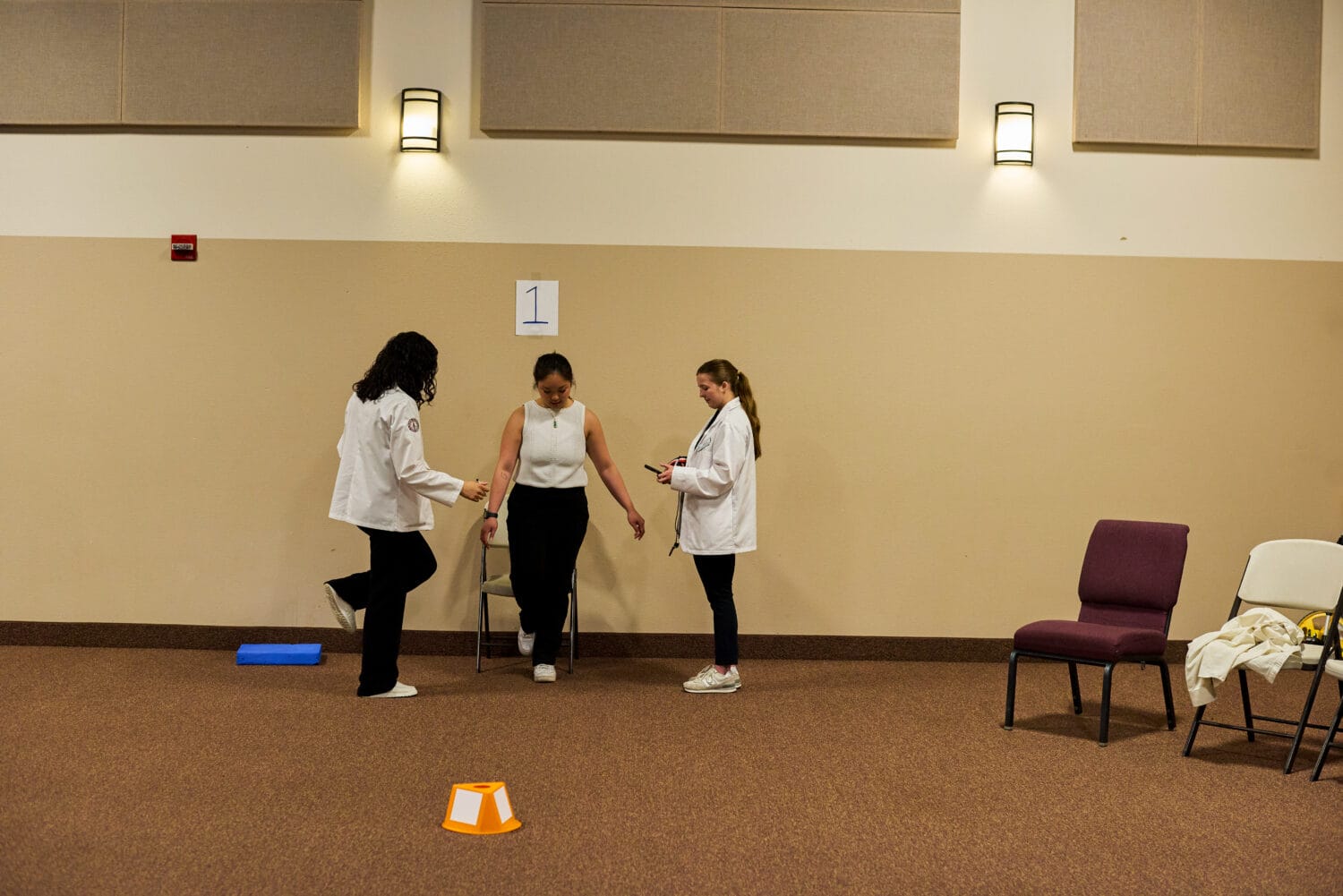 Three women stand near a wall in a room; two in white coats observe as the third performs a balance test next to a chair marked with a “1” sign.