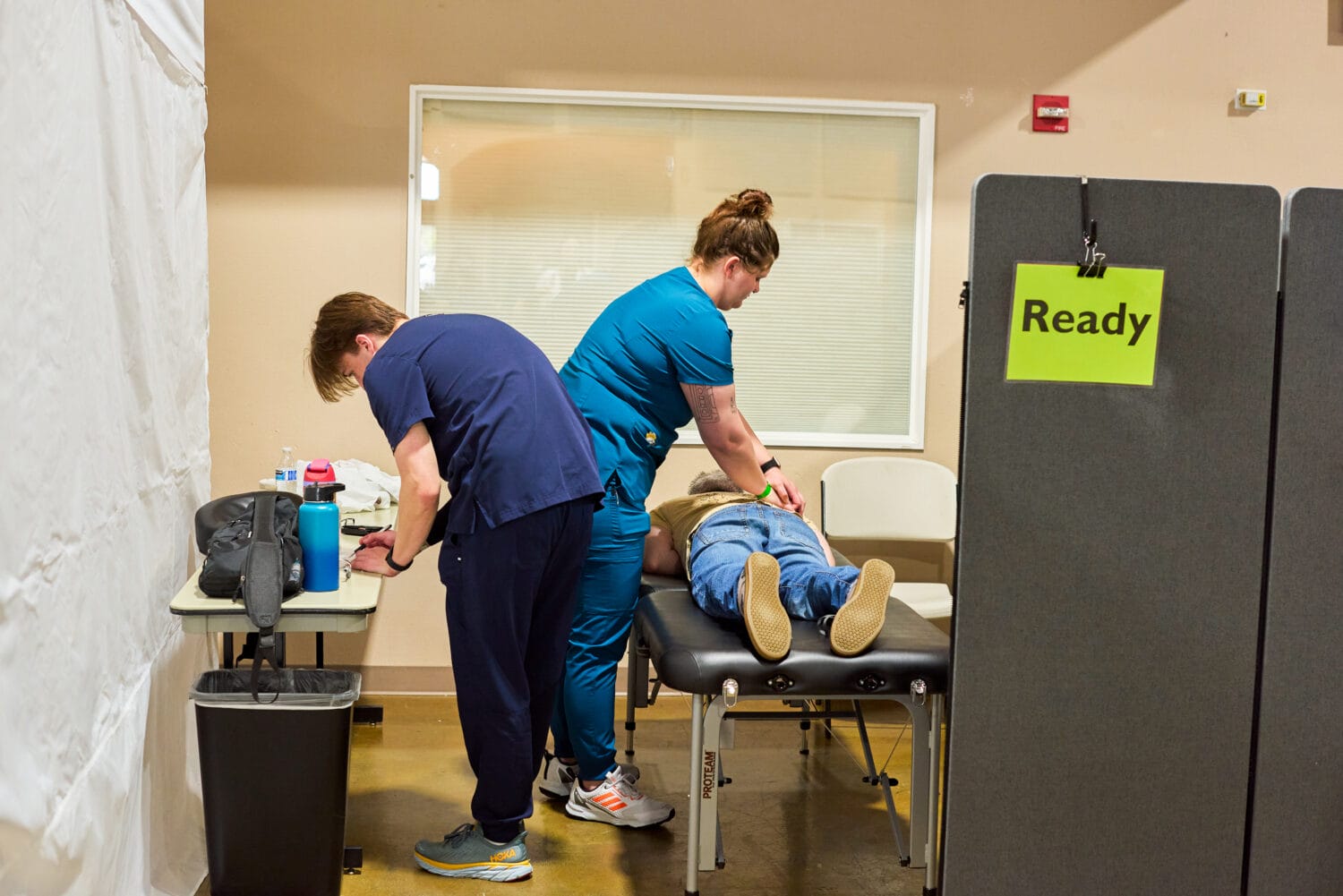 Two healthcare workers attend to a person lying face down on an exam table in a clinic room with a sign labeled "Ready" nearby.
