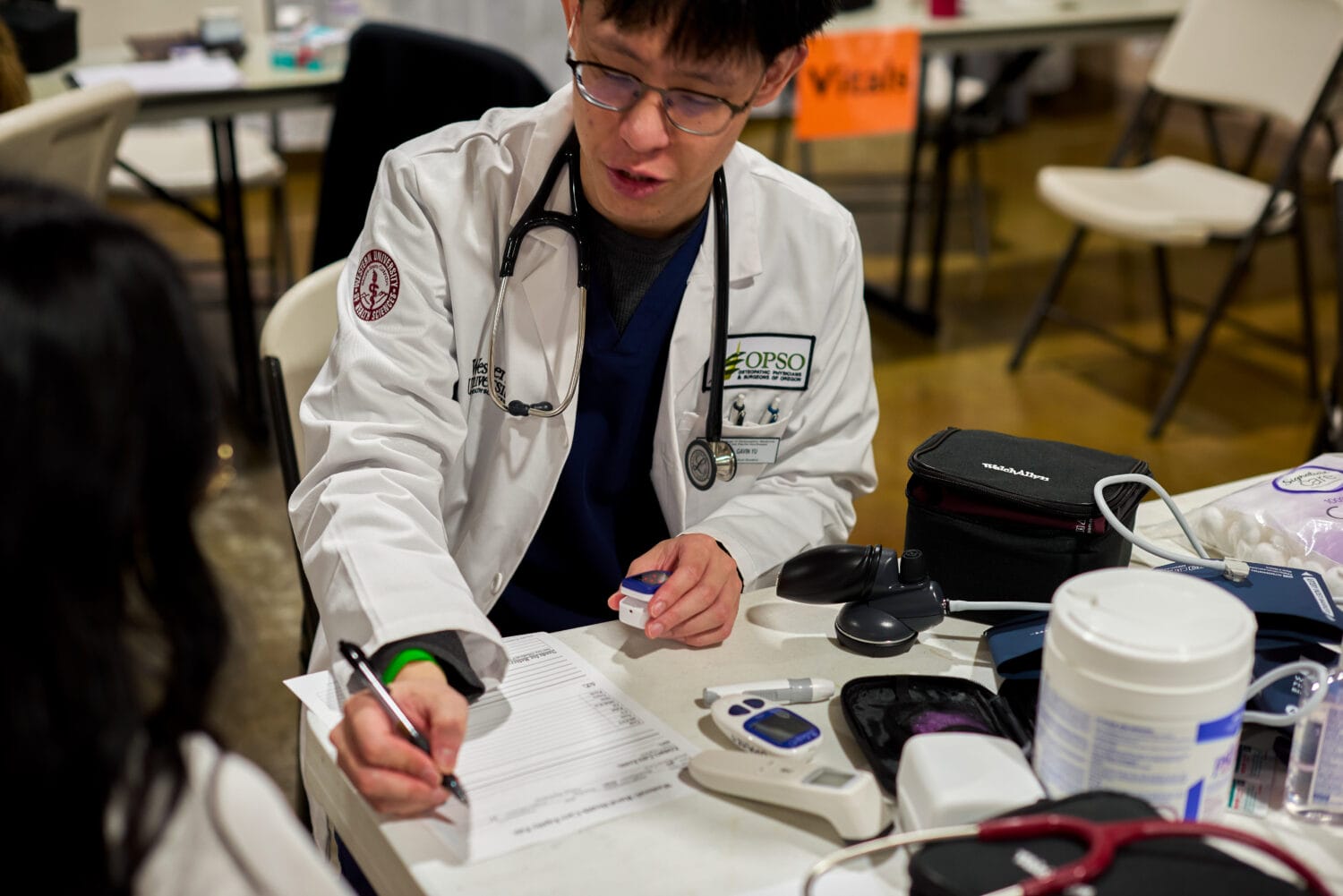 A medical professional in a white coat writes on a form at a table with medical devices, while speaking to a person seated across from him.