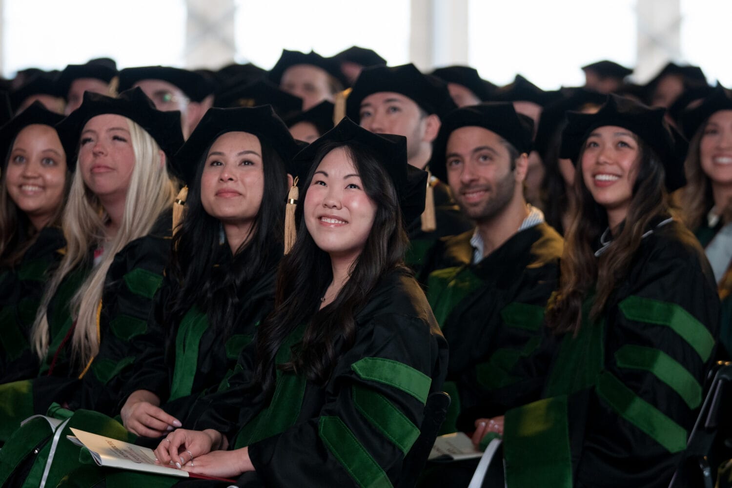 A group of graduates in caps and gowns sit together at a ceremony, looking forward and smiling.