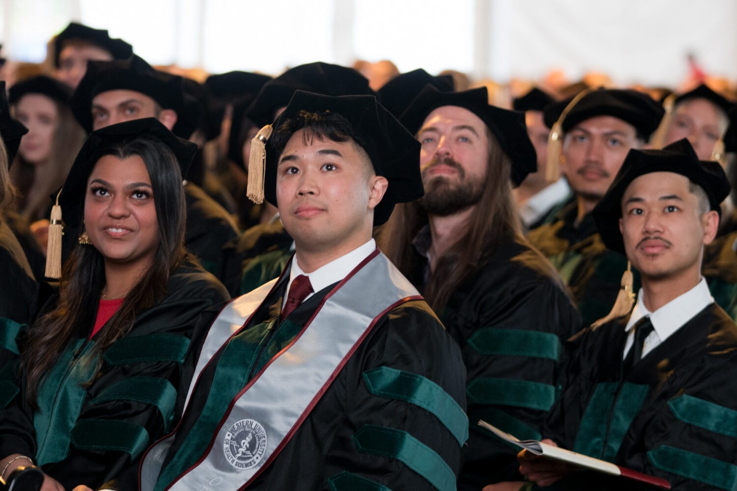 A group of graduates in academic regalia, including caps and gowns, sit in rows during a commencement ceremony, facing forward attentively.