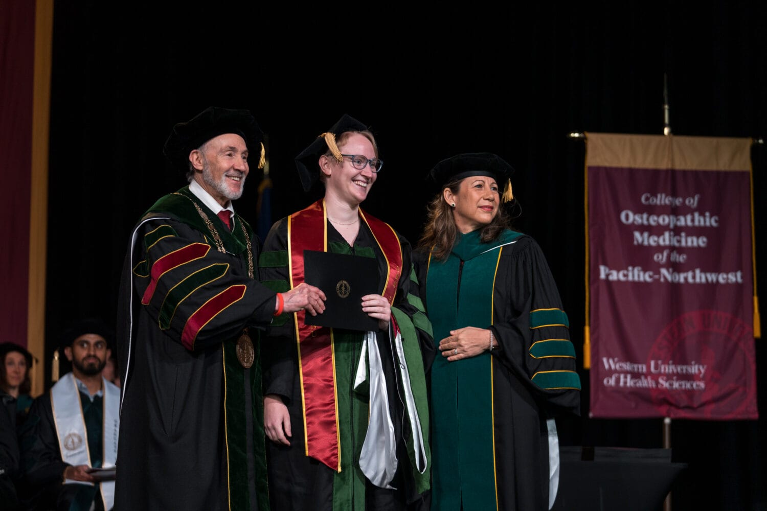 Three people in academic regalia pose on stage; the center person holds a diploma. A banner reads "College of Osteopathic Medicine of the Pacific-Northwest.