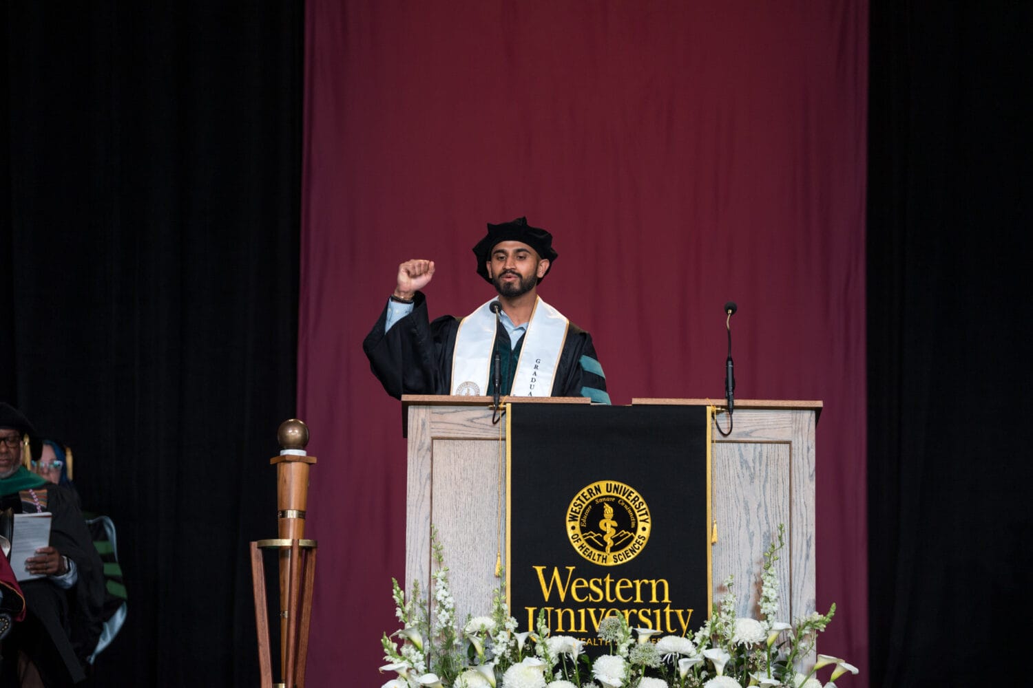 A speaker in graduation attire raises a fist at a podium displaying a "Western University" banner, with flowers in the foreground and a maroon curtain in the background.