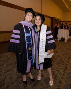 Two women in graduation gowns and caps pose indoors; one wears a "Graduate Speaker" sash. Both are smiling at the camera at what appears to be a graduation event.