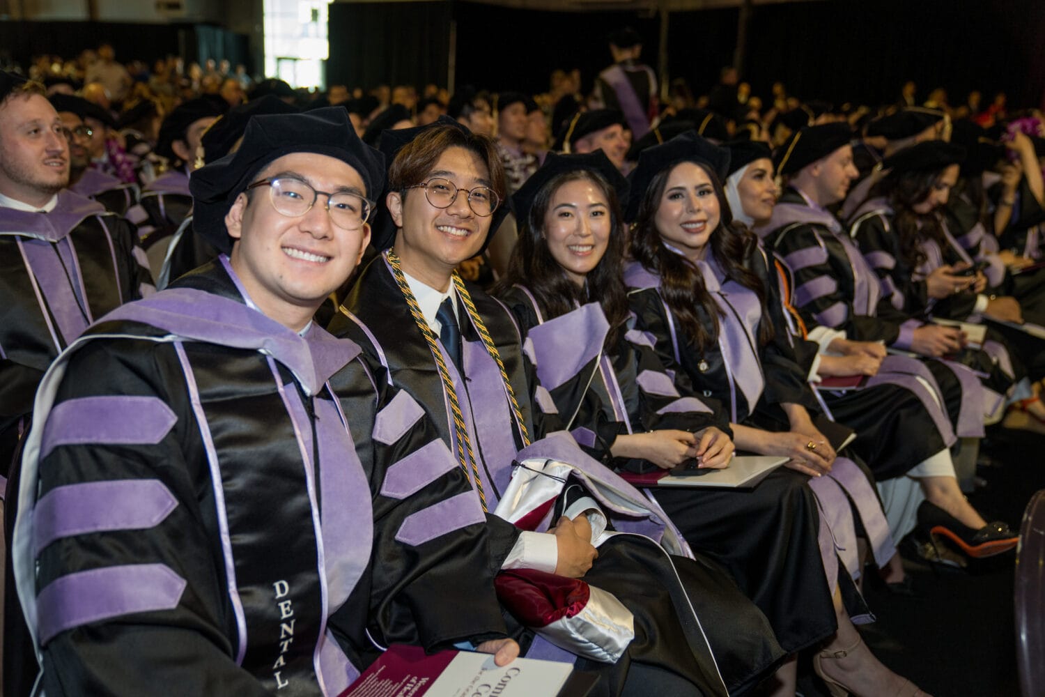 A group of graduates in caps and gowns sit in rows during a graduation ceremony, smiling at the camera.