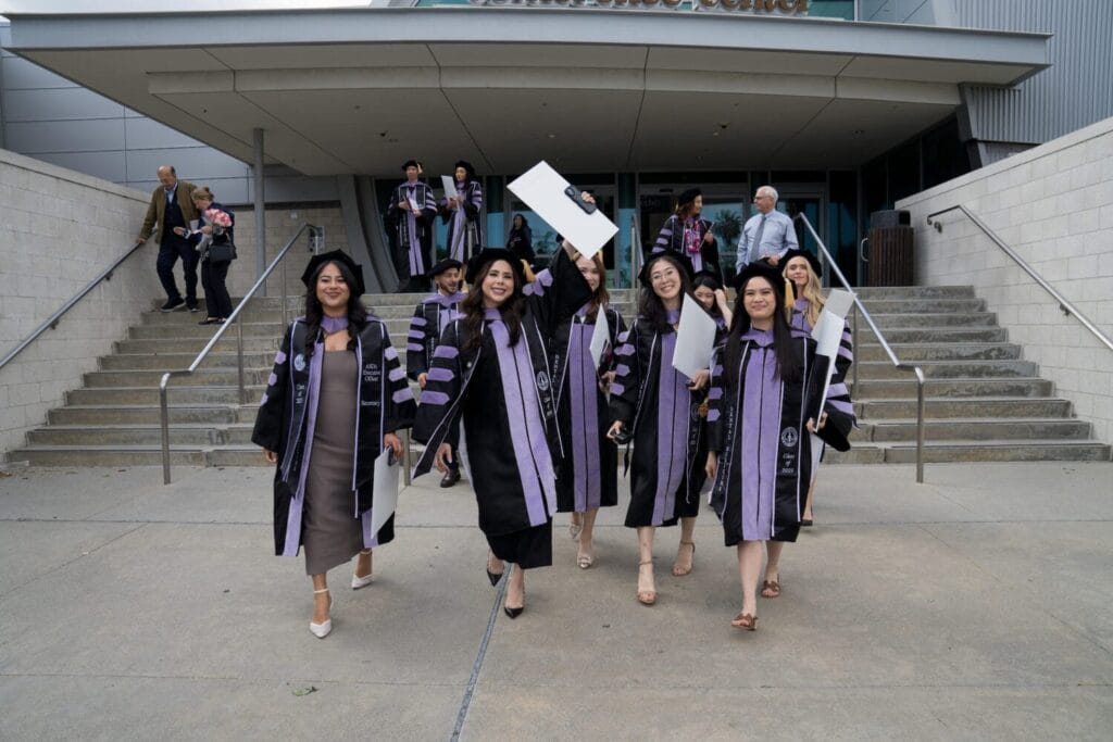 A group of graduates in caps and gowns walk down steps outside a building, some holding diplomas and smiling.