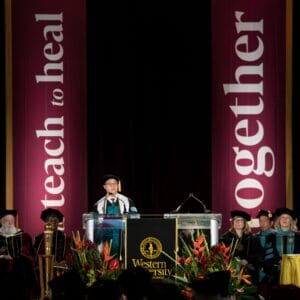 A person speaks at a podium on a stage during a graduation ceremony at Western University of Health Sciences, with faculty seated behind and banners reading "teach to heal" and "together.