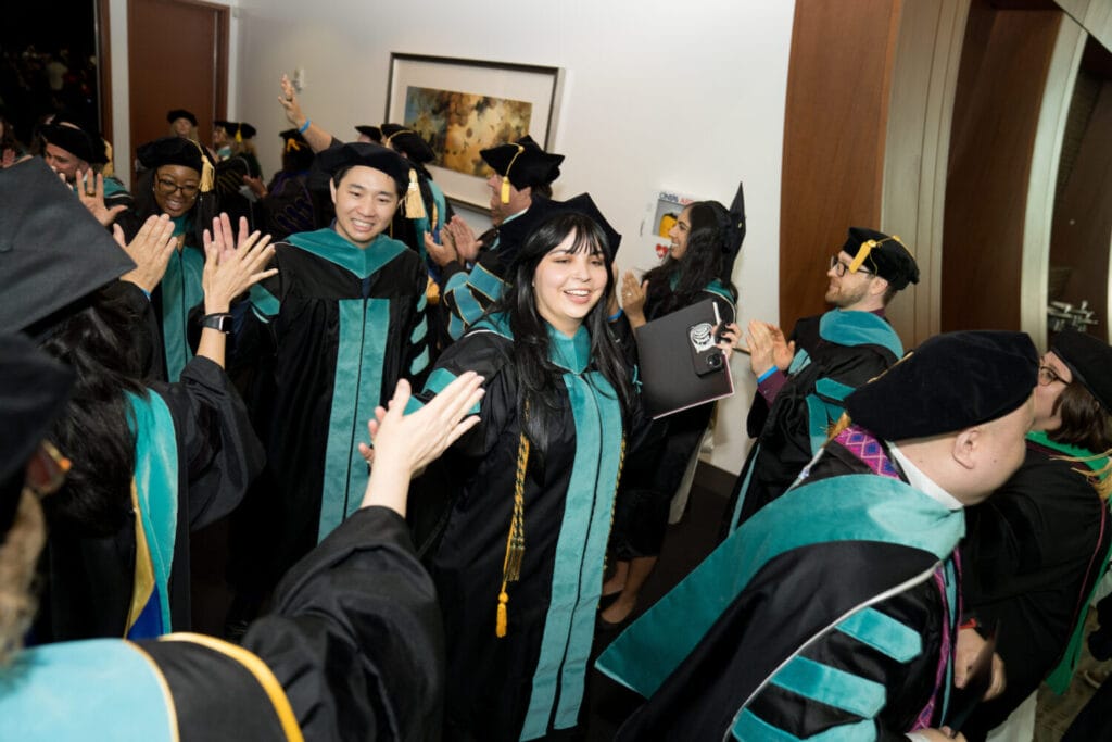 Graduates in academic regalia smile and high-five each other while walking through a hallway during a graduation ceremony.