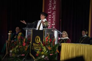 A speaker in academic regalia gestures at a podium labeled "Western University" during a graduation ceremony, with officials seated behind and floral arrangements in front.