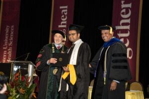 Three men in academic regalia stand on stage at a graduation ceremony. The man in the center holds a diploma while the two others smile for the photo. Banners hang in the background.