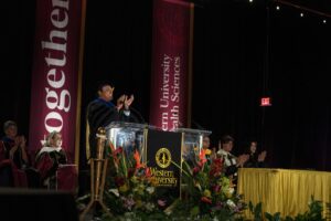 A speaker in academic regalia stands at a podium on stage during a Western University Health Sciences event, with faculty seated and banners in the background.