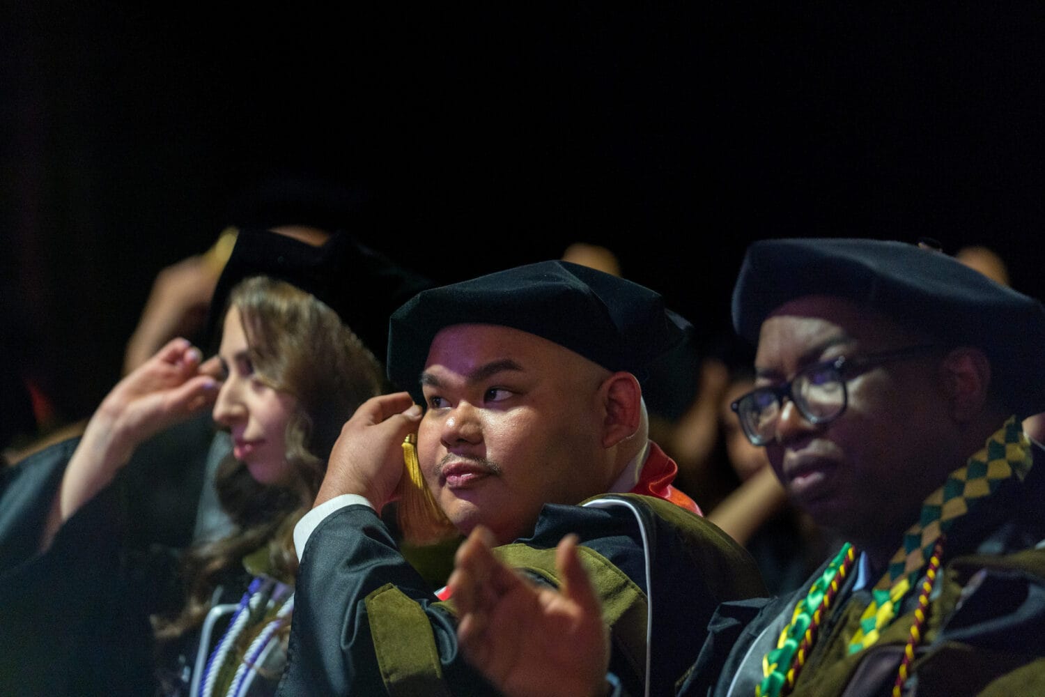 Three graduates in caps and gowns sit in a row at a ceremony, adjusting their tassels and looking ahead.