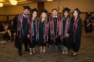 Six graduates in caps and gowns stand in a row and smile for a group photo at an indoor graduation ceremony.