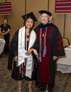 A graduate in cap and gown stands next to a faculty member in academic regalia at an indoor graduation ceremony.