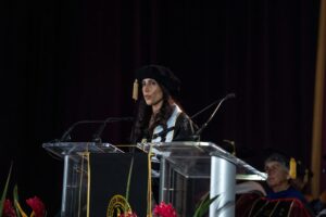 A woman in academic regalia speaks at a podium during a graduation ceremony, with other officials seated behind her on stage.