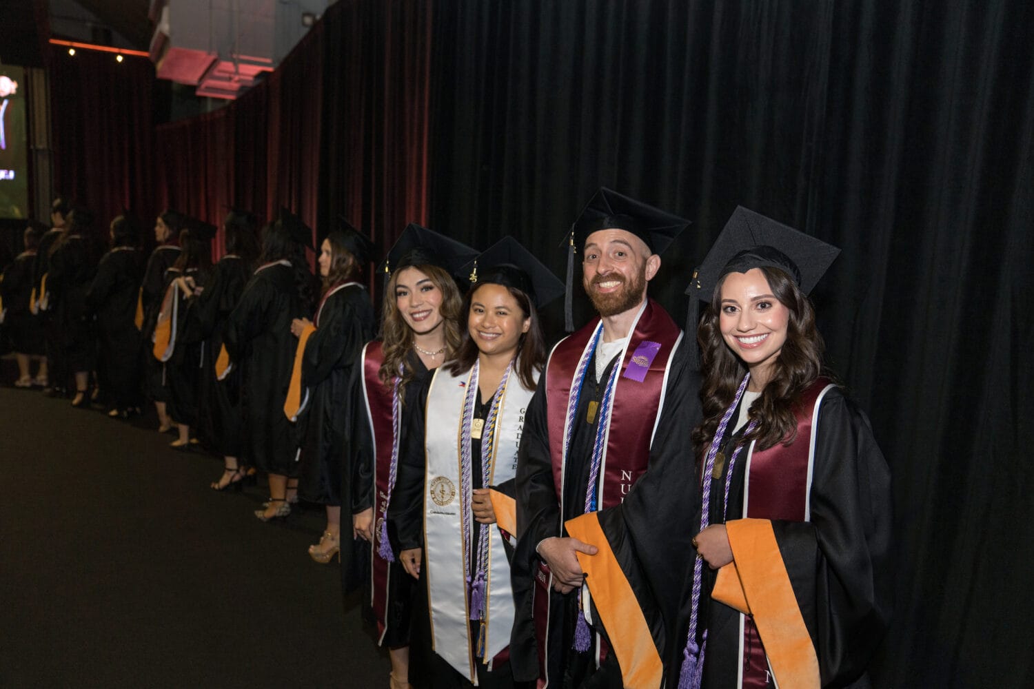 Four graduates in caps and gowns stand smiling in front of a line of other graduates at a graduation ceremony indoors.