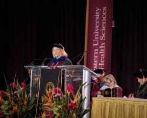 A woman in academic regalia speaks at a podium during a graduation ceremony at Western University of Health Sciences, with other officials seated nearby.
