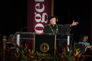 A university official in academic regalia speaks at a podium during a graduation ceremony, with the Western University logo and floral arrangements visible.