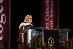 A woman in academic regalia speaks at a podium during a university event, with banners and floral arrangements visible in the background.
