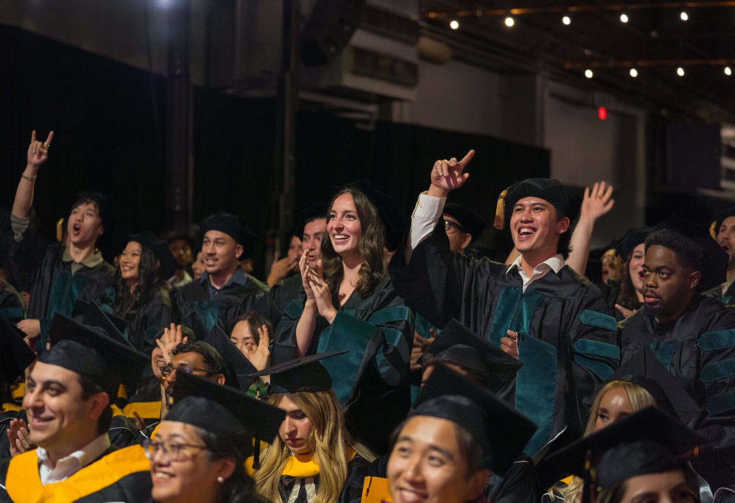 A group of graduates in caps and gowns sit and cheer at an indoor graduation ceremony under string lights.