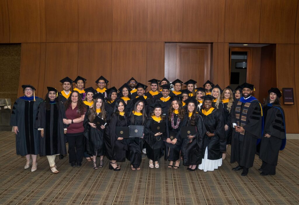 A group of graduates in caps and gowns pose indoors with two faculty members, some holding diplomas, in front of wooden paneling.