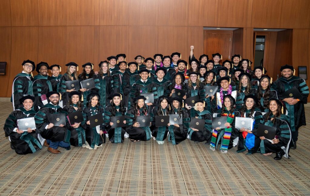 A large group of graduates in caps and gowns poses indoors, holding diplomas and smiling at the camera.