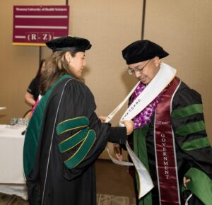 A faculty member in academic regalia places a sash on a graduating student wearing a cap and gown at a graduation event.