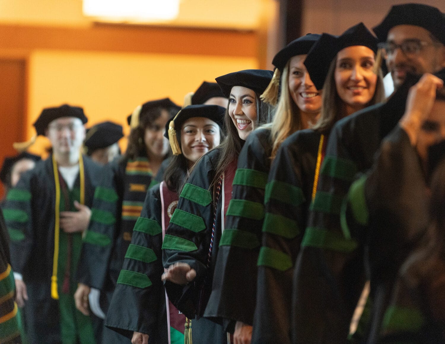 A group of graduates wearing academic caps and gowns stand in a line indoors, smiling and facing forward.