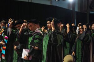 A group of graduates in caps and gowns moves their tassels during a graduation ceremony, holding diplomas and standing in rows.