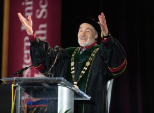 A man in academic regalia stands at a podium with arms raised, smiling, during a formal event.